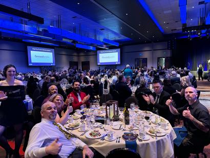 People sitting at round tables in a large banquet hall, with a presentation on screens in the background.