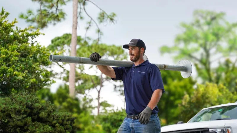 Man in blue shirt carrying long metal pole outdoors, near trees and a white truck.