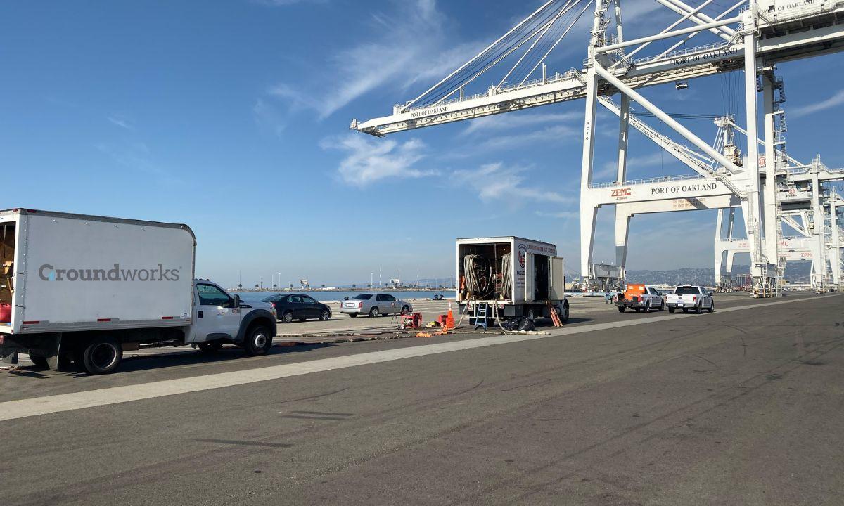 Trucks and equipment near cranes at a pier under a clear sky, with "Port of Oakland" visible on a crane.