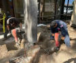 Two workers with jackhammers on a sidewalk, surrounded by construction tools, near a commercial building in daylight.