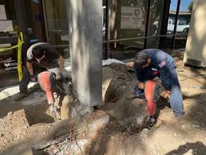 Two workers with jackhammers on a sidewalk, surrounded by construction tools, near a commercial building in daylight.