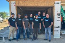 Group of people in front of a partially constructed building, all wearing matching black shirts with green and blue logos.