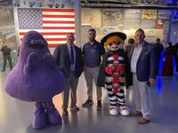 People in mascot costumes and suits pose for a photo at an event with an American flag backdrop.