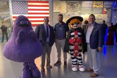 People in mascot costumes and suits pose for a photo at an event with an American flag backdrop.