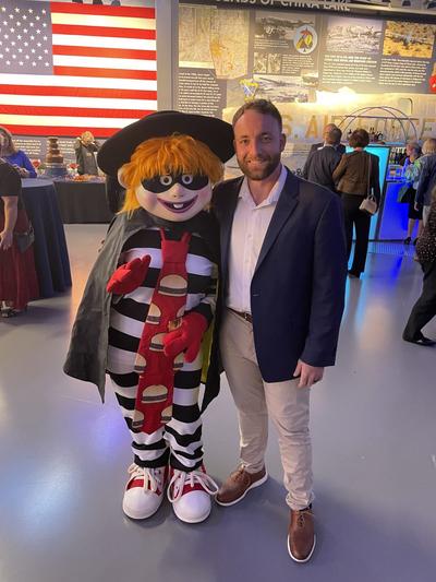 A man poses with a costumed character in a museum setting with an American flag in the background.