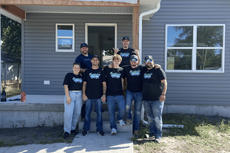 Group of people in matching shirts stand smiling in front of a newly built small gray house.