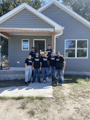 Group of people in matching shirts stand smiling in front of a newly built small gray house.