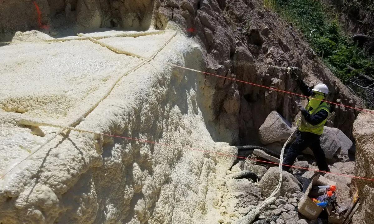 Worker in safety gear sprays yellow foam onto a rocky surface near a cliff, surrounded by equipment.