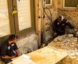 Two workers in uniforms repair a multi-family residential foundation, tools and debris scattered around, near a stone wall.
