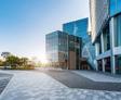Modern office building with glass facade, adjacent trees, and a sunlit courtyard with a paved walkway.