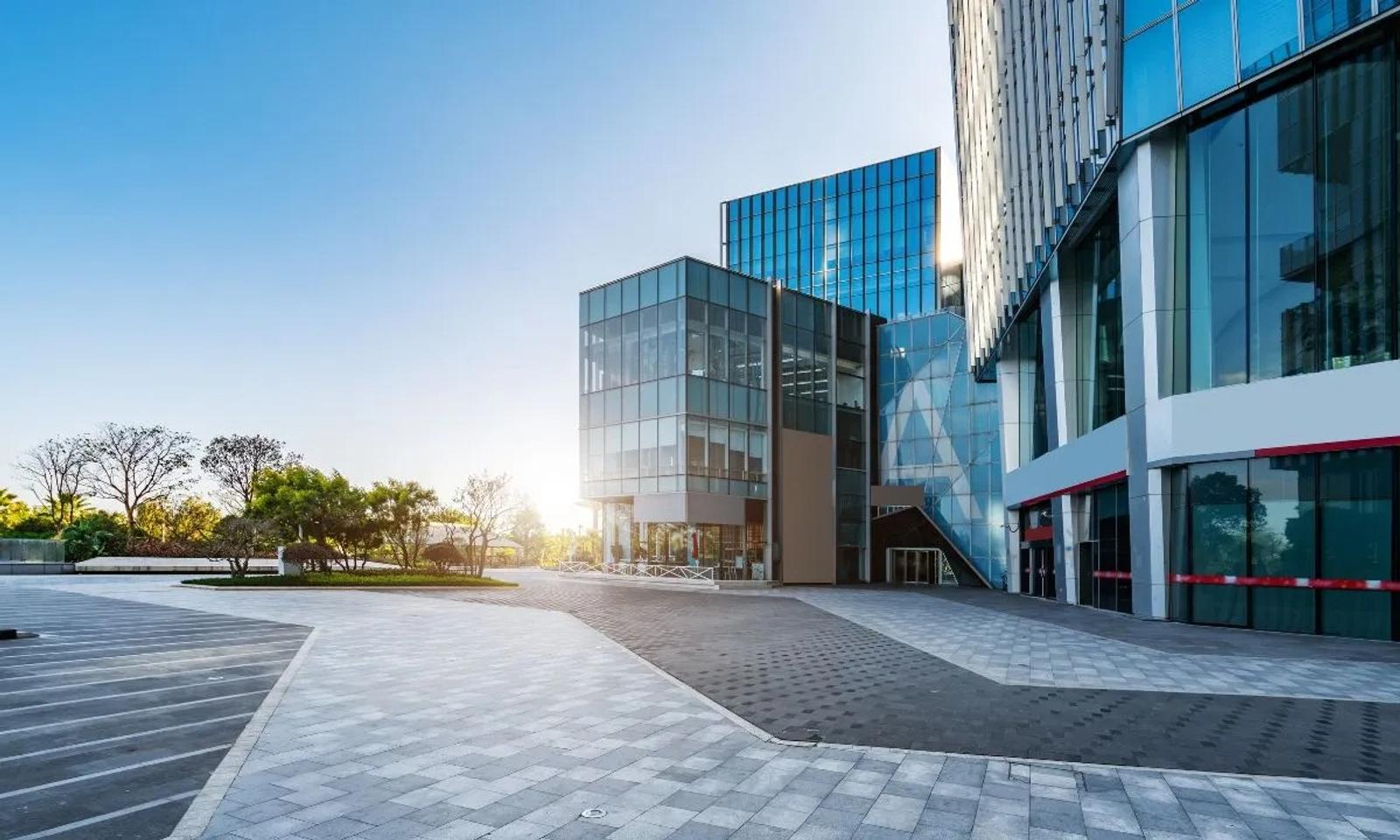 Modern office building with glass facade, adjacent trees, and a sunlit courtyard with a paved walkway.