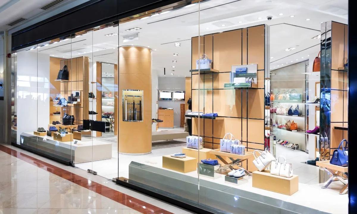 Storefront with large glass windows displaying various handbags and shoes on shelves and pedestals inside a shop.