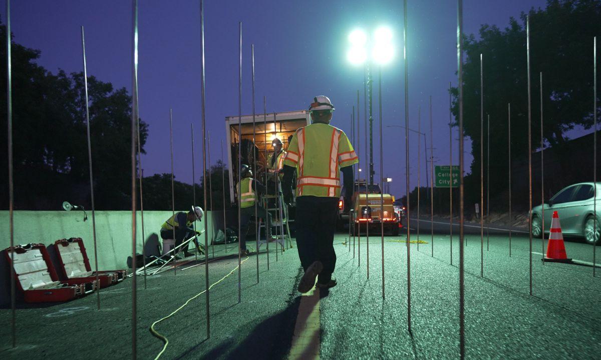 Workers in safety vests set up equipment on a road at night, surrounded by tall poles and bright lights.