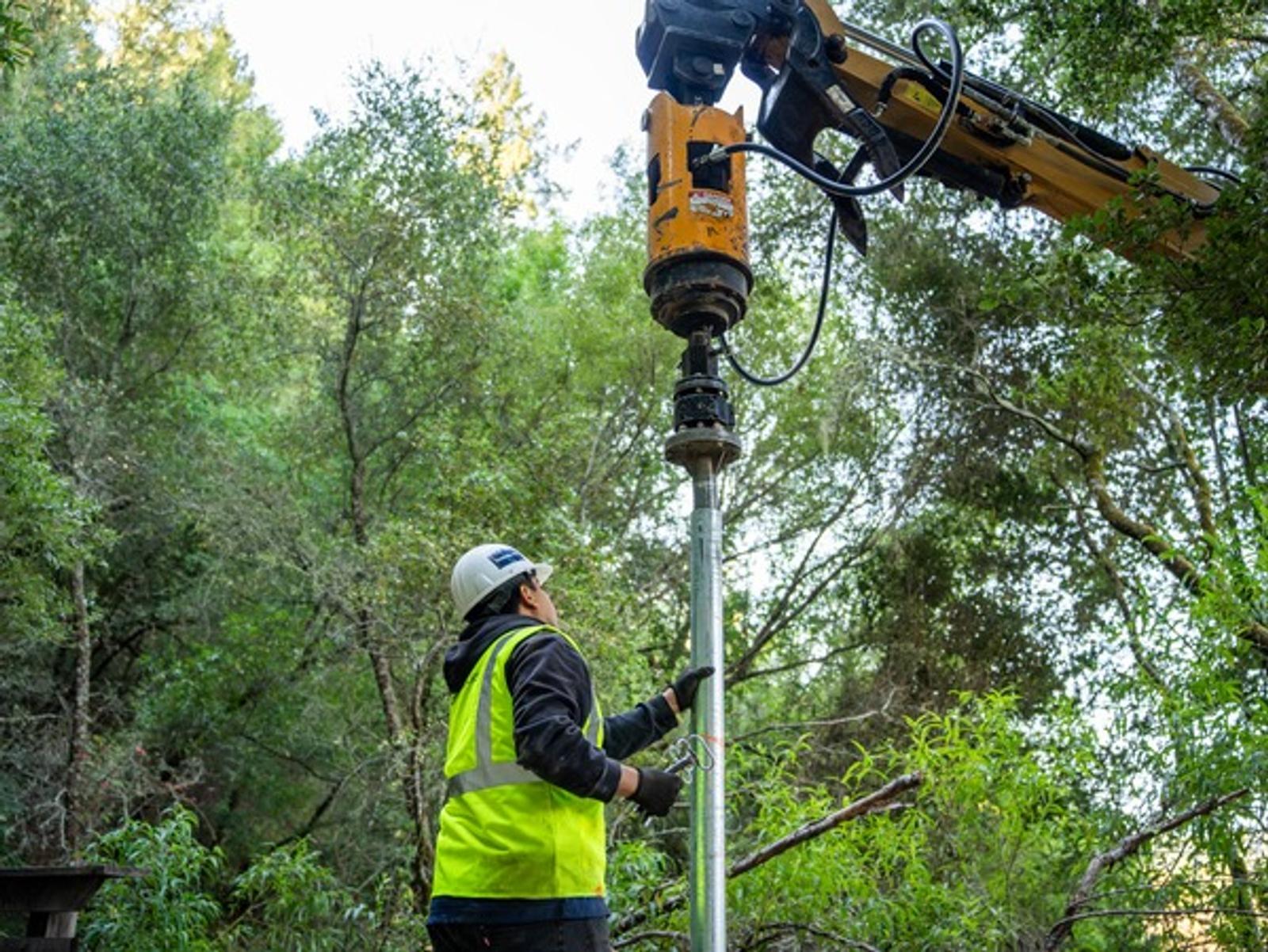 Worker in a hard hat operates a large drill in a forested area, surrounded by green trees.