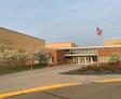 School building with a U.S. flag, brick and beige facade, surrounding trees, and a paved walkway.