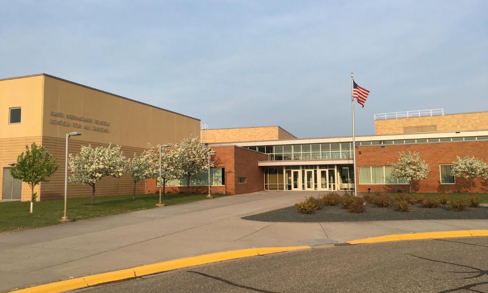 School building with a U.S. flag, brick and beige facade, surrounding trees, and a paved walkway.