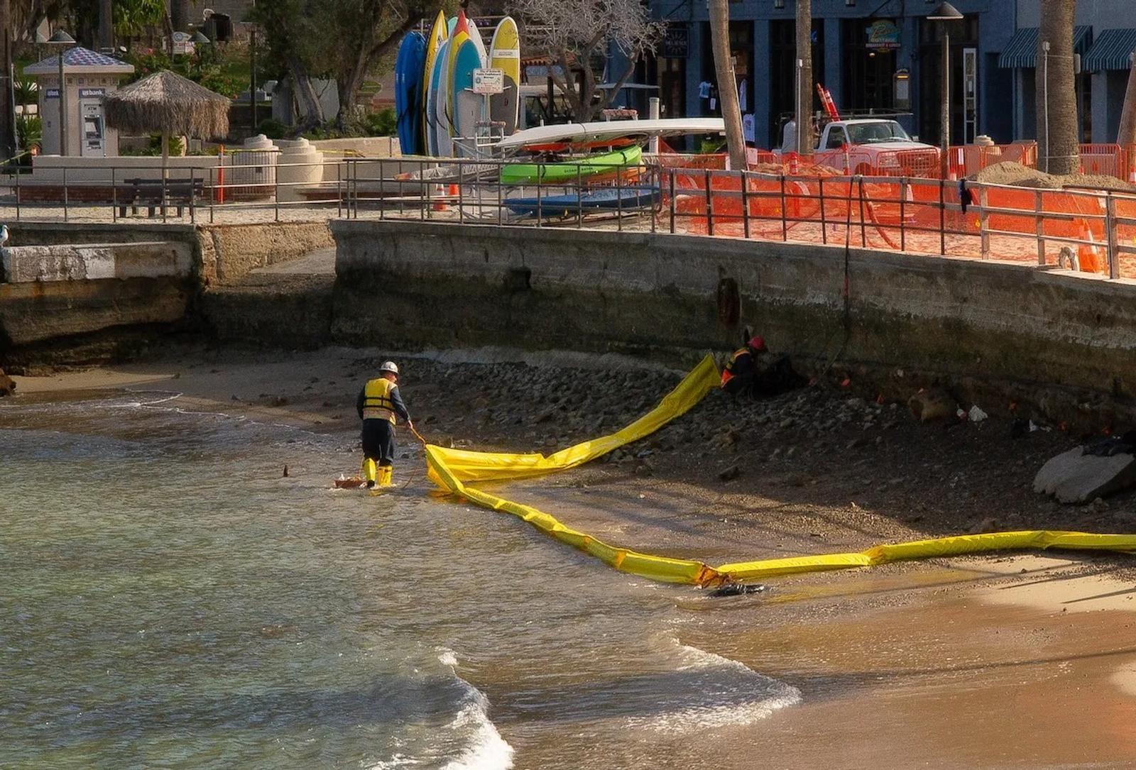 Workers in safety gear place a yellow barrier along the sandy shoreline, focusing on seawall stabilization near the construction fencing.