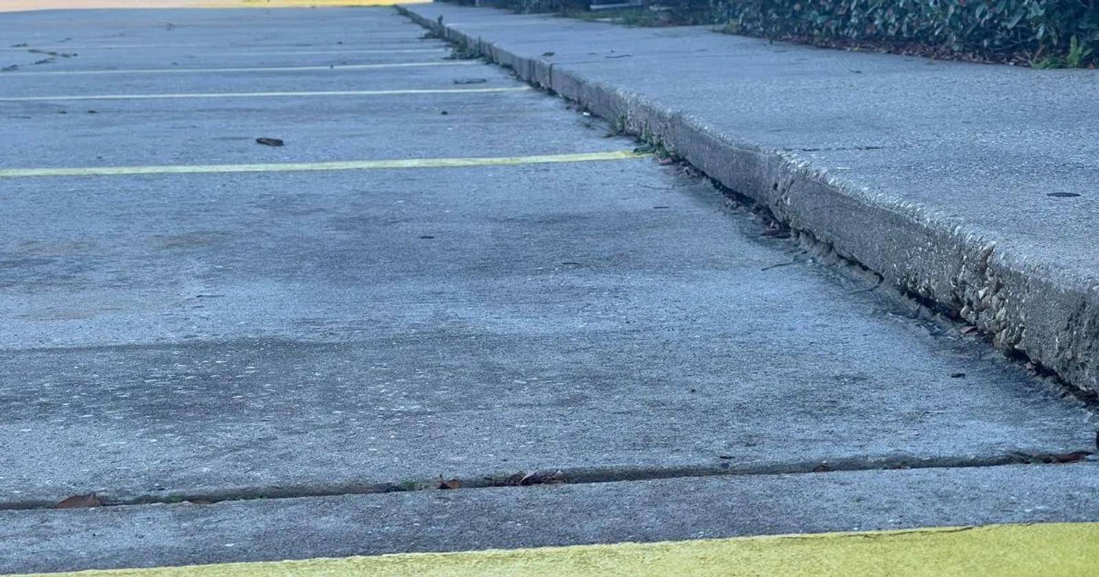 Empty parking lot with cracked pavement needing leveling, yellow lines, and a sidewalk lined with greenery.