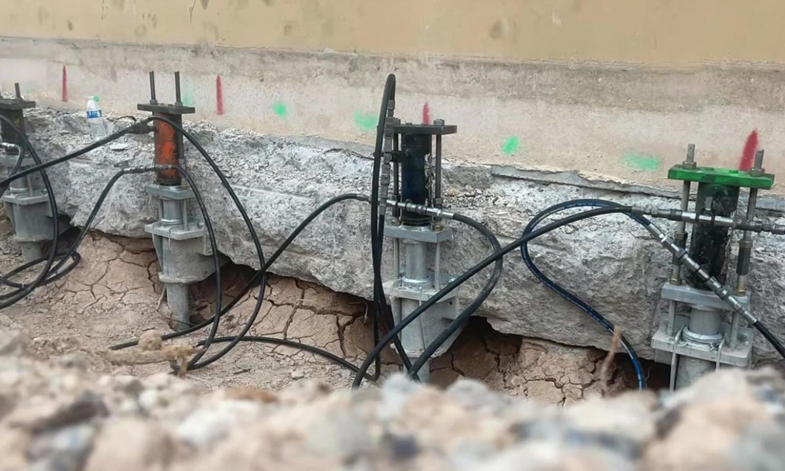 Hydraulic jacks installed under a cracked foundation wall with exposed cables on a construction site.