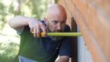 A man measures the side of a wooden building with a yellow tape measure for crawl space encapsulation outdoors.