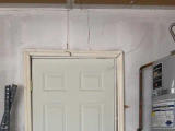 White door with a damaged frame and cracks in the wall above, next to a water heater in a garage.