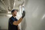 A man wearing gloves and a cap installs a vapor barrier on a basement wall, pressing it firmly.
