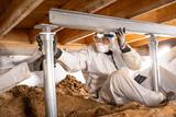 A worker in protective gear installs metal supports in a crawl space under a wooden floor.