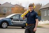 Smiling worker holding a level tool stands by a Groundworks vehicle, ready for geotextile reinforcement in a suburban neighborhood.