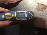 Hand holding a digital moisture meter pressed against a wooden surface, displaying a reading on the screen.