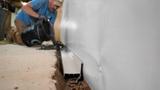 A person installs a drainage system along a basement wall, pouring material into a trench on the floor.