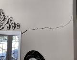 A long crack runs across a beige wall, above a door and a clock, near decorative metal wall art.
