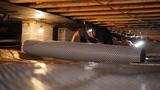 Person wearing a headlamp works in a crawl space, unrolling geotextile reinforcement material on the ground.