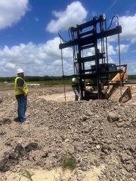 Construction workers in safety gear operate heavy machinery, focusing on soil stabilization for new developments at a rocky site under a partly cloudy sky.