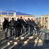 Group of people in warm clothing standing inside a partially constructed wooden building under a clear blue sky.