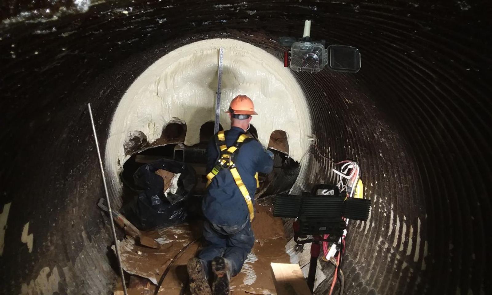A worker in safety gear meticulously maintains defense infrastructure inside a large pipe, surrounded by essential tools and equipment.