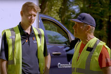 Two people in safety vests stand talking near a vehicle with "Groundworks" on the door. Trees are visible in the background.