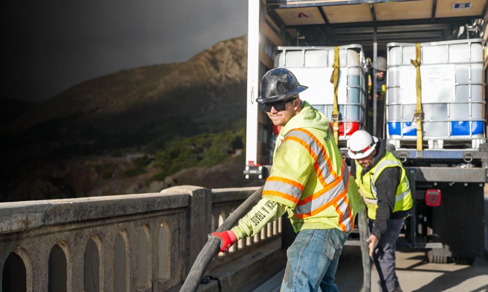 Construction workers wearing safety gear unload equipment from a truck on a bridge, with hills in the background.