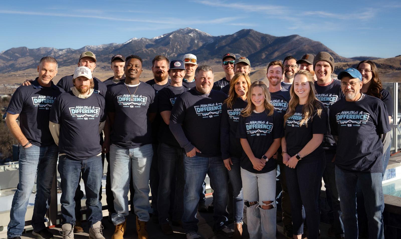 A group of people wearing matching T-shirts poses outdoors with mountains in the background.