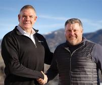 Two men shaking hands outdoors, with mountains in the background under a clear blue sky.