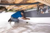 Man installing a vapor barrier on the wall of a crawl space with insulation overhead and pipes nearby.
