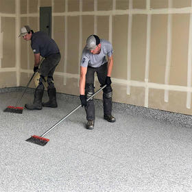 Two workers in gray shirts and gloves apply a speckled finish to a room's floor using long-handled brushes.