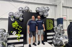 Two people in matching shirts stand on a podium surrounded by silver balloons and tennis ball decorations.