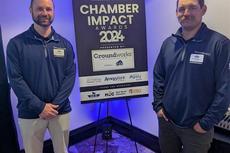 Two people standing beside a "Chamber Impact Awards 2024" sign with sponsor logos in a conference room.