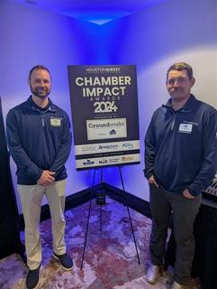 Two people standing beside a "Chamber Impact Awards 2024" sign with sponsor logos in a conference room.