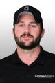 A man with a beard wears a black Groundworks polo shirt and a black cap, posing against a gray background.