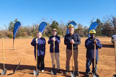 Four people with shovels at a groundbreaking ceremony on a sunny day, standing on a dirt area with banners.