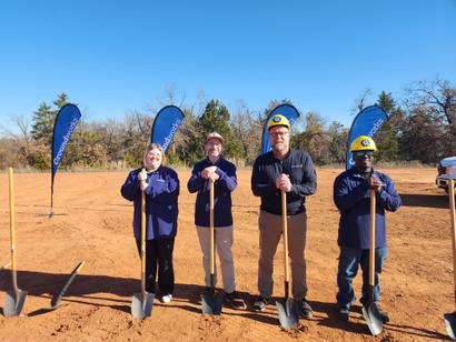 Four people with shovels at a groundbreaking ceremony on a sunny day, standing on a dirt area with banners.