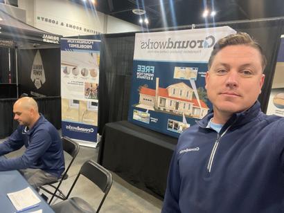 Two men at a trade show booth featuring home construction services and roofing displays.