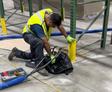 Worker in a warehouse kneeling, fixing shelving with tools, wearing a yellow safety vest and gloves.