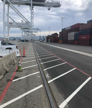 Port with cranes, shipping containers, and a rail track; empty parking spaces line the road.